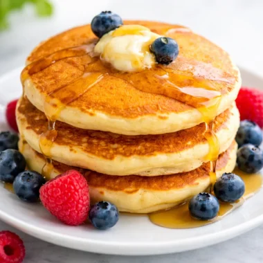Fluffy pancakes stacked on a plate with syrup and berries, showcasing a delicious pancake recipe.